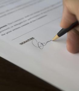 A hand signs a formal contract with a pen on a wooden desk.